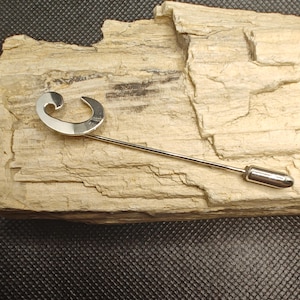 May include: A silver-toned lapel pin with a curved, crescent-shaped top and a long, straight pin. The pin is displayed on a light brown, textured surface with a dark background. The pin's design is simple and elegant.
