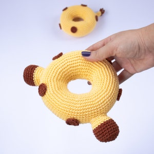 May include: A crocheted yellow donut-shaped toy with brown spots. The toy is held in a hand.