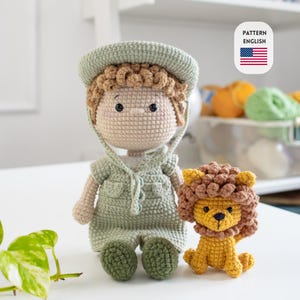 Pode incluir: Duas bonecas de crochê: um menino com uma roupa de safari verde e um leão amarelo. A boneca do menino tem cabelo castanho claro e um chapéu a condizer. O leão tem uma juba castanha e está sentado. A imagem inclui uma pequena bandeira americana e as palavras "PATTERN ENGLISH".