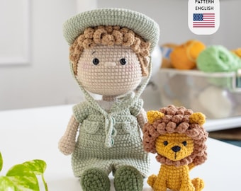 May include: Two crocheted dolls: a boy in a green safari outfit and a yellow lion. The boy doll has light brown hair and a matching hat. The lion has a brown mane and is sitting. The image includes a small American flag and the words "PATTERN ENGLISH".