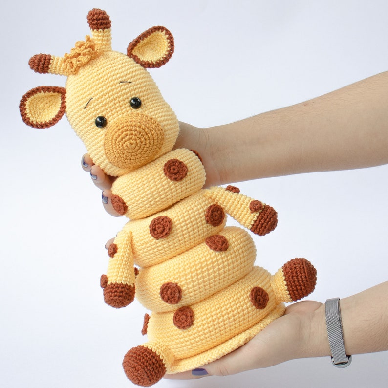 May include: A crocheted giraffe toy with a stack of rings. The giraffe is yellow with brown spots and has a brown nose and eyes. The rings are yellow with brown spots.