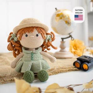 Pode incluir: Boneca de crochê feita à mão com cabelo encaracolado ruivo, vestindo um vestido verde claro e um chapéu bege. A boneca tem bochechas rosadas e está sentada em uma superfície de serapilheira. O texto "PATTERN ENGLISH" também é visível.
