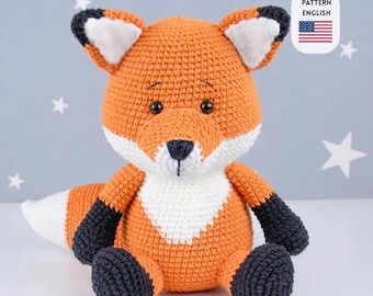 May include: A crocheted fox toy in orange, white, and black. The fox has a white chest and inner ears, black accents on the paws and ears, and a white-tipped tail. The text "PATTERN ENGLISH" and an American flag are in the upper right corner.