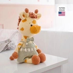 Pode incluir: Uma boneca girafa de crochê com corpo creme, manchas marrons e focinho amarelo. Ela usa um macacão cinza e uma gravata borboleta. A boneca está sentada em uma superfície branca. A imagem inclui o texto "PATTERN ENGLISH" e uma bandeira dos EUA.