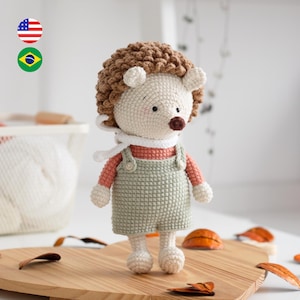 Pode incluir: Uma boneca de ouriço de crochê feita à mão com uma cabeça espinhosa castanha clara, um rosto branco e um nariz castanho. Ela usa uma camisa cor de pêssego, macacão verde e um cachecol branco. A boneca está em uma tábua de madeira.