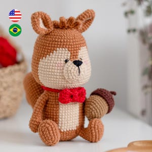 Pode incluir: Um esquilo de pelúcia feito em crochê com fio marrom e creme. O esquilo usa uma gravata borboleta vermelha no pescoço e segura uma bolota de crochê marrom em uma das mãos. Possui olhos pretos bordados, nariz preto e bochechas rosadas.