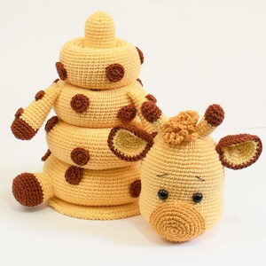 May include: A crocheted giraffe toy with brown spots. The giraffe is made up of four stacked rings, with the top ring forming the head. The giraffe has large brown eyes and a small brown nose.