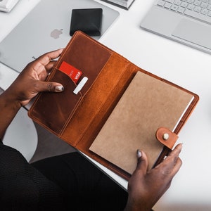 Puede incluir: Un cuaderno de cuero marrón con una ranura para tarjetas y un cierre de botón dorado. El cuaderno está abierto y las páginas están en blanco.
