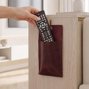 May include: A dark burgundy leather remote control holder hanging from a small brass hook on the side of a light wood end table, containing a black television remote control with numerous buttons and a small red indicator light.