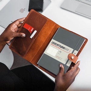 May include: A brown leather notebook cover with a red credit card and a pen in the pocket. The cover is open to reveal a grey notebook with the text "LEUCHTTURM1917" on the front.