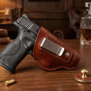 May include: A black semi-automatic handgun partially inserted into a brown leather holster featuring a silver metal belt clip. Two brass-colored cartridges are positioned on the surface near the base of the handgun grip.