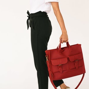 May include: A red leather tote bag with two front pockets and buckle closures. The bag has a shoulder strap and top handles. The person is wearing a white t-shirt, black pants, and white sneakers.