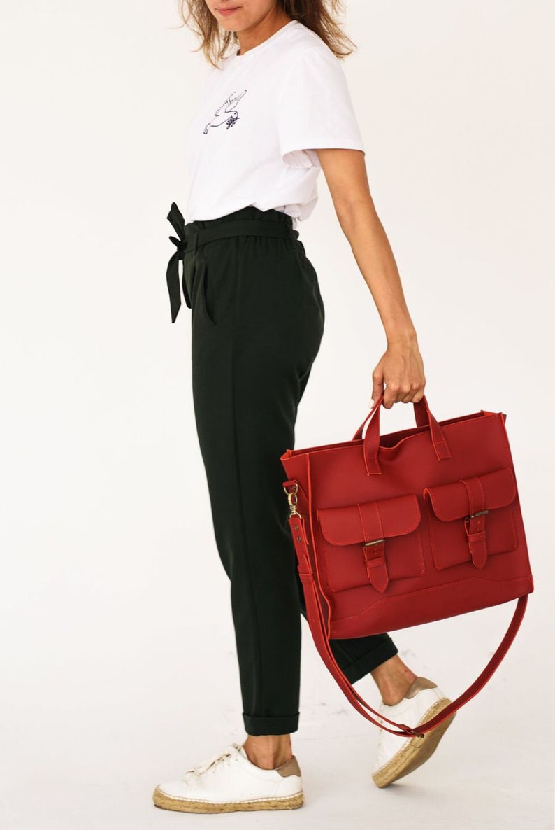 May include: A red leather tote bag with two front pockets and buckle closures. The bag has a shoulder strap and top handles. The person is wearing a white t-shirt, black pants, and white sneakers.