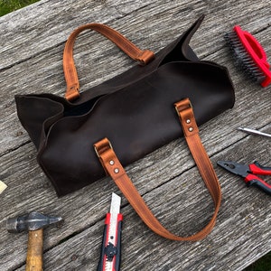 Pode incluir: Uma bolsa tote de couro castanho escuro com duas al&ccedil;as de couro castanho claro fixadas por rebites met&aacute;licos. A bolsa tem uma abertura superior e uma base retangular. Est&aacute; rodeada por v&aacute;rias ferramentas, incluindo um martelo, x-ato e alicates.