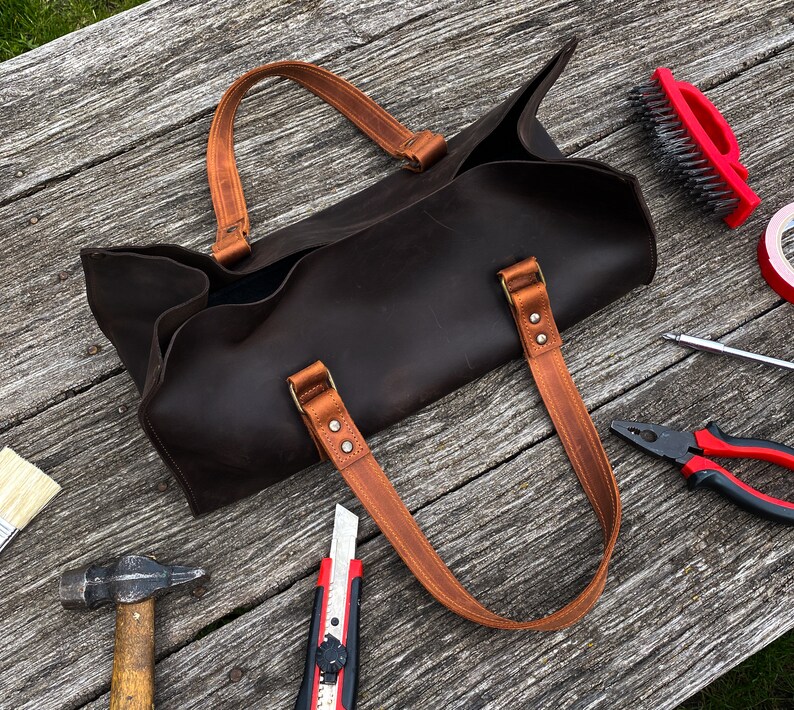Pode incluir: Uma bolsa tote de couro castanho escuro com duas al&ccedil;as de couro castanho claro fixadas por rebites met&aacute;licos. A bolsa tem uma abertura superior e uma base retangular. Est&aacute; rodeada por v&aacute;rias ferramentas, incluindo um martelo, x-ato e alicates.
