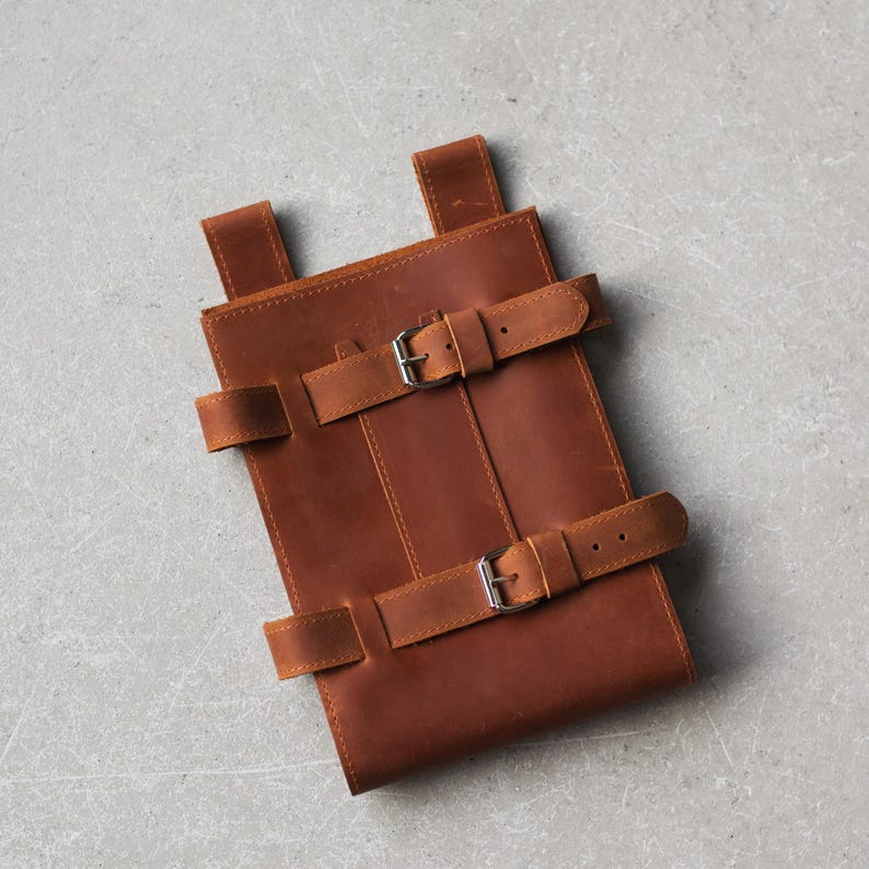 May include: A brown leather carrying case with two buckles and straps. The case is made of a thick, durable leather and has a simple, classic design.