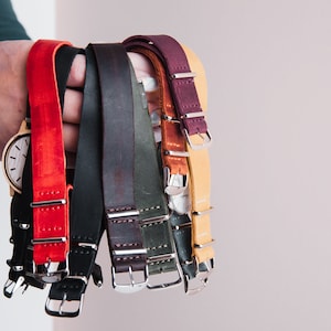 May include: A collection of eight leather watch straps in various colors, including red, black, brown, green, and tan. The straps have silver buckles and are arranged in a hand.
