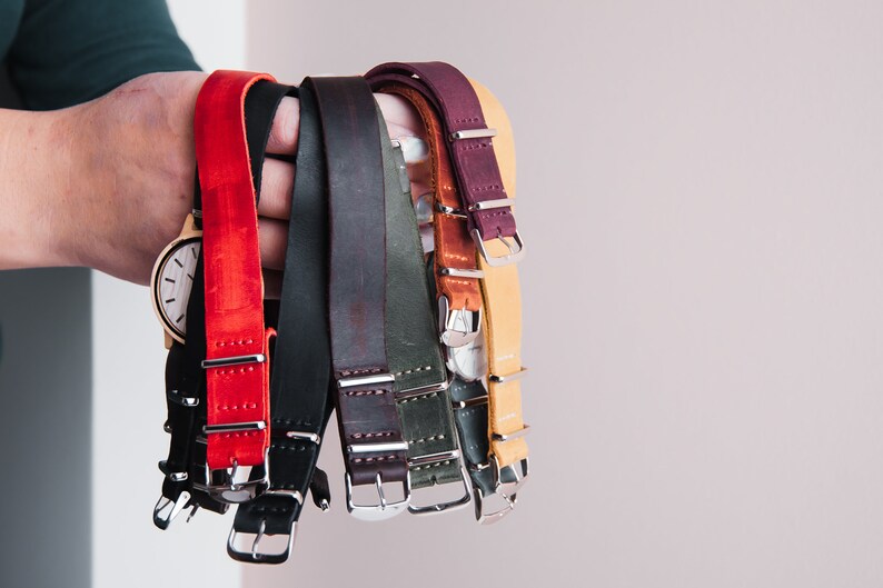 May include: A collection of eight leather watch straps in various colors, including red, black, brown, green, and tan. The straps have silver buckles and are arranged in a hand.