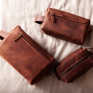 May include: Three brown leather wash bags of different sizes. Each bag features a zip closure and a handle. The leather has a distressed appearance. The bags are arranged on a neutral-coloured fabric.