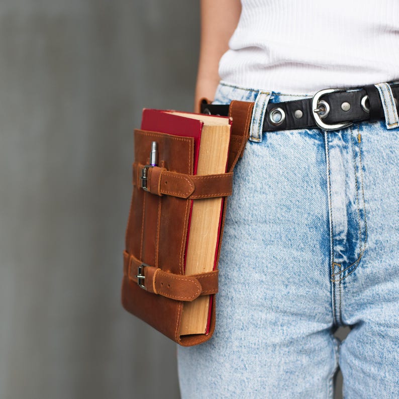 May include: A brown leather book holder with a strap and buckles. The holder is worn on a person's hip and holds a book with a red cover.