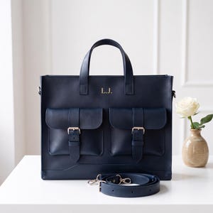 Könnte beinhalten: Eine marineblaue Aktentasche aus Leder mit zwei Vordertaschen mit Schnallenverschlüssen, zwei Tragegriffen und einem abnehmbaren Schultergurt. Auf der Vorderseite der Tasche sind die goldfarbenen Initialen L.J. zwischen den Griffen eingeprägt.