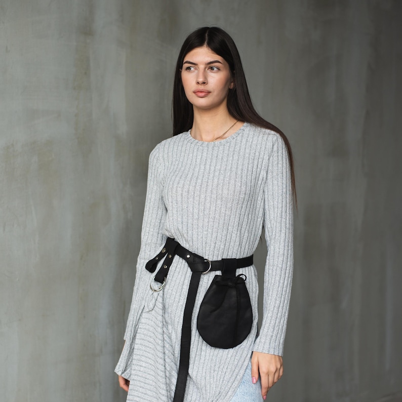 May include: A woman wearing a gray ribbed sweater dress with a black belt and a small black pouch attached. The belt has silver hardware and the pouch has a drawstring closure.