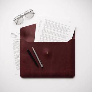 May include: A dark burgundy leather document sleeve with a single gold-tone stud closure. The sleeve contains white printed papers. A pair of silver-tone wire-frame glasses, a black pen, and a silver pen are positioned next to the document sleeve.