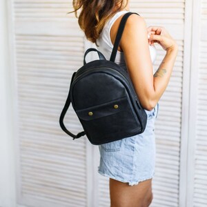 May include: A black leather backpack with a flap closure and a small pocket on the front. The backpack has brass hardware and a distressed look.