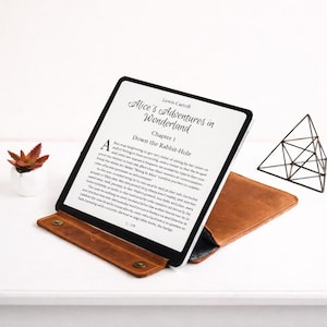 May include: An e-reader in a brown leather case, displaying "Alice's Adventures in Wonderland". The screen shows the book's text. A small succulent in a white pot and a geometric metal sculpture are also visible on a white surface.