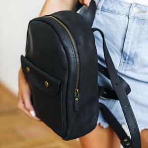 May include: A black leather backpack with a gold zipper and a flap pocket with a brass button. The backpack has adjustable straps.