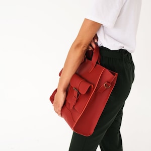 May include: A red leather tote bag with a flap pocket and a buckle closure. The bag has a top handle and a shoulder strap attachment. The bag is held by a person wearing a white shirt and dark green pants.