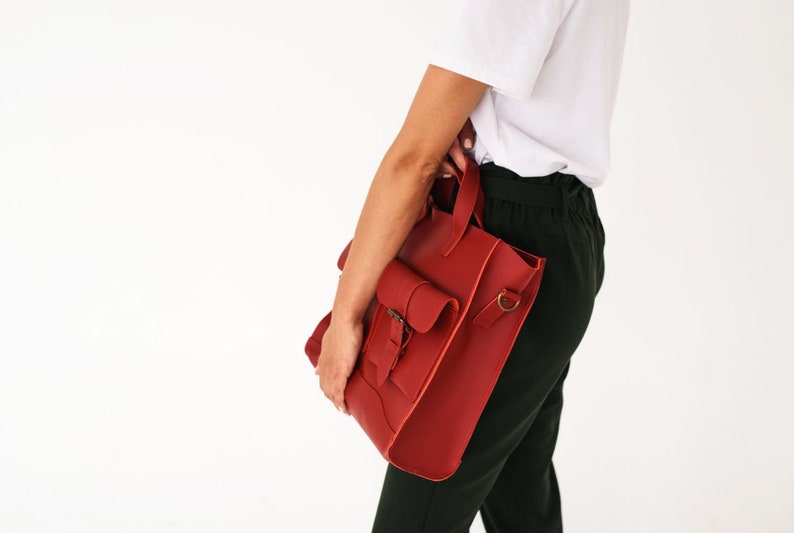 May include: A red leather tote bag with a flap pocket and a buckle closure. The bag has a top handle and a shoulder strap attachment. The bag is held by a person wearing a white shirt and dark green pants.