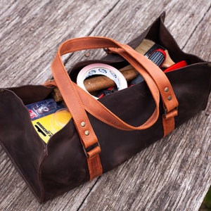 Pode incluir: Uma bolsa de ferramentas retangular castanho-escura com al&ccedil;as de couro castanho-claro e rebites de lat&atilde;o. A bolsa est&aacute; cheia com v&aacute;rios itens, incluindo um rolo de fita branca, uma escova com cabo de madeira e uma embalagem amarela com texto.