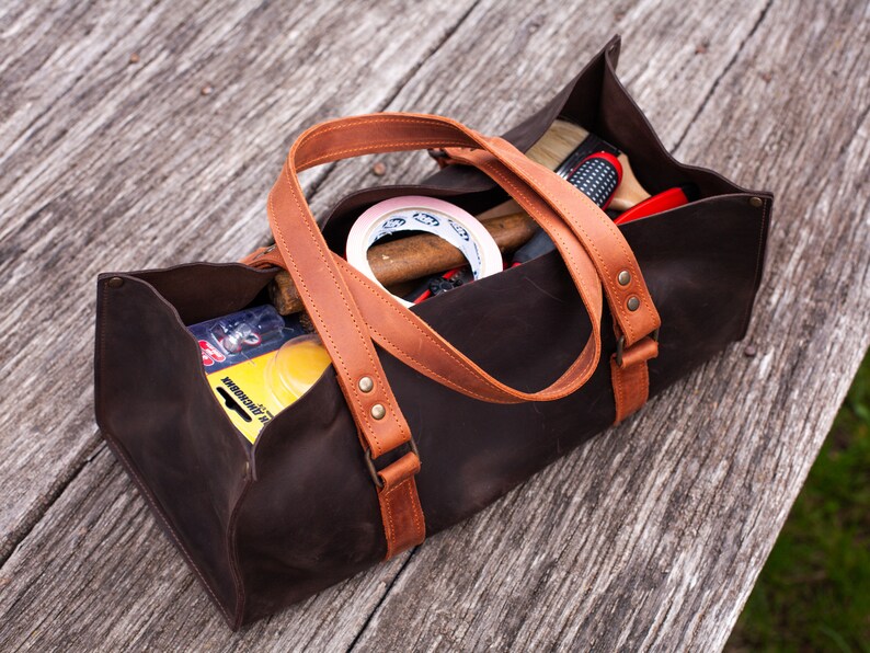 Pode incluir: Uma bolsa de ferramentas retangular castanho-escura com al&ccedil;as de couro castanho-claro e rebites de lat&atilde;o. A bolsa est&aacute; cheia com v&aacute;rios itens, incluindo um rolo de fita branca, uma escova com cabo de madeira e uma embalagem amarela com texto.