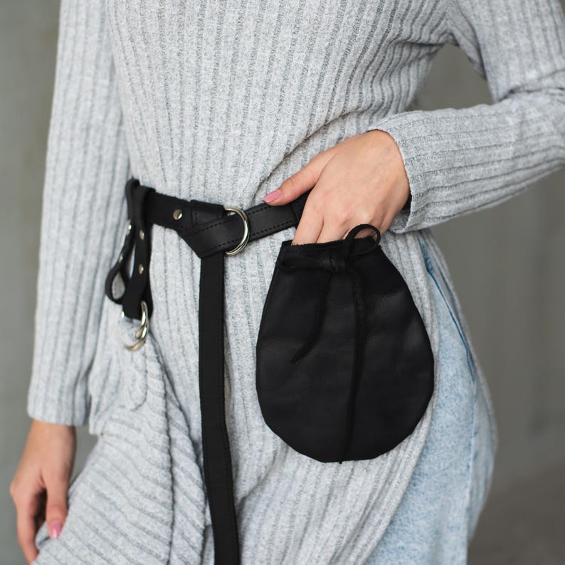 May include: A black leather belt with a silver buckle and a small black pouch attached. The belt is worn over a gray sweater and blue jeans.