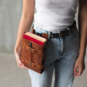 May include: A brown leather book holder with a red book inside. The holder is attached to a black belt with silver buckles. The belt is worn around the waist of a person wearing blue jeans.