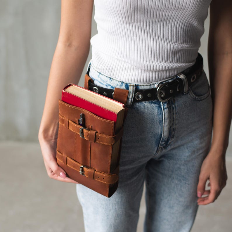 May include: A brown leather book holder with a red book inside. The holder is attached to a black belt with silver buckles. The belt is worn around the waist of a person wearing blue jeans.