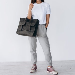 May include: A dark gray leather tote bag with multiple front pockets and buckle closures. The bag is held by a person wearing a white t-shirt and gray pants. The bag appears to be made of a durable material.