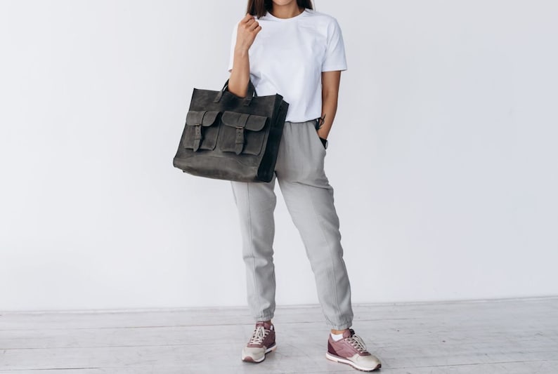 May include: A dark gray leather tote bag with multiple front pockets and buckle closures. The bag is held by a person wearing a white t-shirt and gray pants. The bag appears to be made of a durable material.