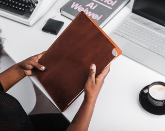 Könnte beinhalten: Eine Person hält ein braunes Ledernotizbuch mit weißem Rand. Das Notizbuch liegt auf einem weißen Schreibtisch mit einem Laptop, einem Buch und einer Tasse Kaffee.