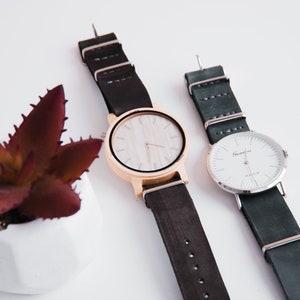 May include: Two wristwatches with black leather bands. The watch on the left has a wooden face with a white dial and gold-colored hands. The watch on the right has a silver face with a white dial and silver hands. The text "Grenville" is printed on the face of the watch on the right.