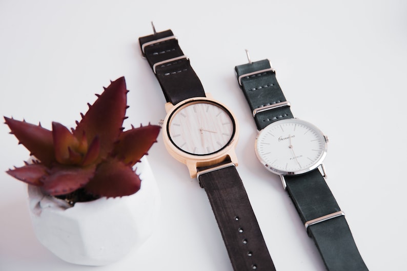 May include: Two wristwatches with black leather bands. The watch on the left has a wooden face with a white dial and gold-colored hands. The watch on the right has a silver face with a white dial and silver hands. The text "Grenville" is printed on the face of the watch on the right.