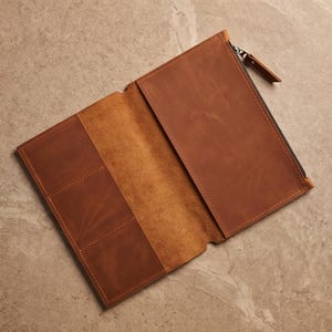 Pode incluir: Uma carteira de couro castanho-claro com acabamento exterior liso e interior texturizado. A carteira inclui um compartimento com fecho de correr no lado direito e três ranhuras verticais para cartões no lado esquerdo, com costuras visíveis.