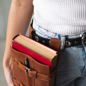 May include: A brown leather book holder with a red and cream book inside. The holder has a buckle and strap closure. The holder is worn on a person's hip with a black belt.