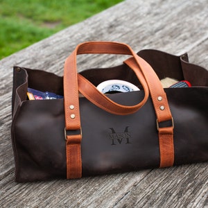 Pode incluir: Uma bolsa de ferramentas de couro castanho escuro com al&ccedil;as de couro bege, rebites cor de lat&atilde;o e o nome MASON gravado na frente. A bolsa retangular aberta cont&eacute;m v&aacute;rios itens, incluindo um rolo de fita branca e uma pequena escova.