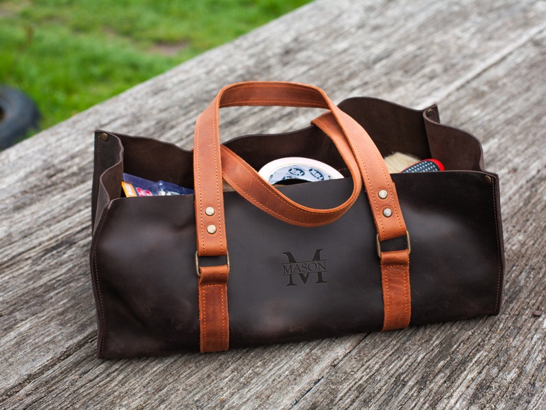 Pode incluir: Uma bolsa de ferramentas de couro castanho escuro com al&ccedil;as de couro bege, rebites cor de lat&atilde;o e o nome MASON gravado na frente. A bolsa retangular aberta cont&eacute;m v&aacute;rios itens, incluindo um rolo de fita branca e uma pequena escova.