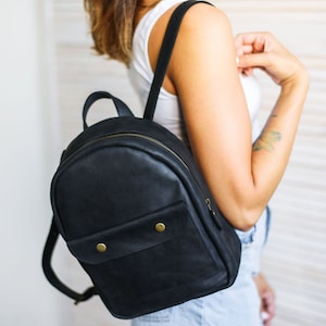 May include: A black leather backpack with a front pocket and gold-toned hardware. The backpack has a rounded shape and a zipper closure.