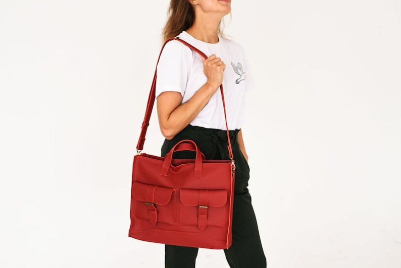 May include: A red leather satchel with two front pockets and a shoulder strap. The bag has a top handle and a rectangular shape. The bag is being carried over the shoulder. The bag is a solid red color.