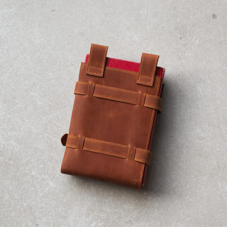 May include: A brown leather journal with a red interior and multiple straps. The journal is closed and lying flat on a gray surface.