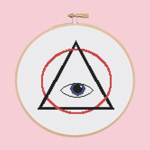 May include: A white embroidery hoop with a black triangle and red circle containing a blue eye. The design is a stylized all-seeing eye.